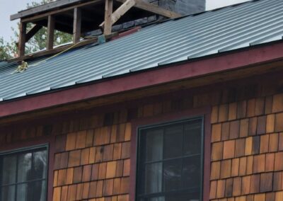 Close-up of a wooden shingle house with dark-framed windows and a newly installed metal roof featuring a chimney with stone base and stainless steel chimney pipe, showcasing property management maintenance and roofing upgrade.