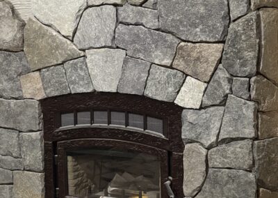 Stone fireplace with dark metal framed glass door insert, surrounded by irregularly shaped gray and beige natural stones, part of an interior home renovation project.