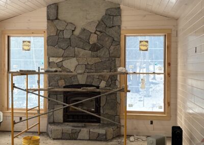 Stone fireplace under construction between two windows in a wood-paneled room with construction scaffolding, tools, and materials, illustrating home improvement and interior renovation in progress.
