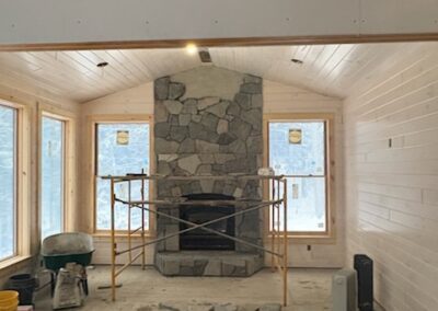 Interior of a room under construction featuring a stone fireplace surrounded by scaffolding, wood-paneled walls and ceiling, large windows, and construction materials scattered on the floor, representing Excelsior Property Management renovation project.