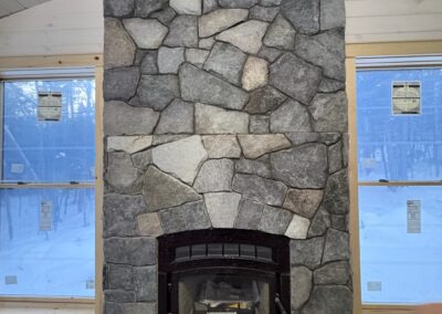 Stone fireplace with black metal wood stove insert in a room under construction, flanked by two large windows showing a snowy outdoor landscape, highlighting rustic interior design and property management renovation.