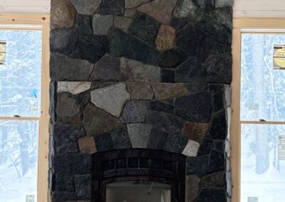 Rustic stone fireplace with dark metal insert surrounded by large natural stones, set between two windows in a snowy cabin interior under construction, showcasing handcrafted masonry for cozy property management spaces.