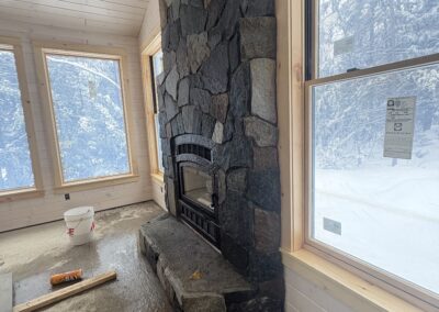 Stone fireplace under construction inside a wood-paneled room with large windows showing a snowy outdoor landscape, part of Excelsior Property Management's winter-ready home projects.