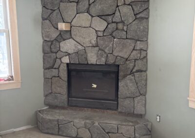 stone fireplace with irregular gray stones and a black metal firebox insert set in an unfinished room with green walls and bare floor during home renovation