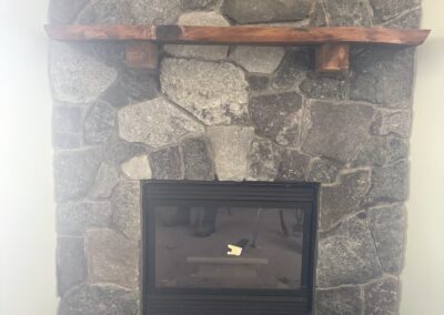 Stone fireplace with large irregular gray stones and a wooden mantel shelf, featuring a built-in black glass-fronted firebox, part of Excelsior Property Management home interior improvements.