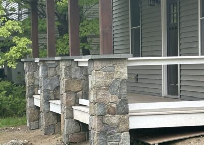Stone pillar supports with wooden beams on the front porch of a residential home featuring gray siding and a black door, illustrating durable exterior construction for property management and home improvement.