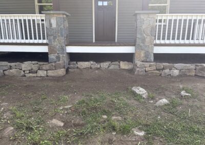 Stone pillars and a low stone wall bordering the front porch of a residential property with bare soil and sparse grass in the foreground, highlighting exterior stonework and landscaping features for Excelsior Property Management.