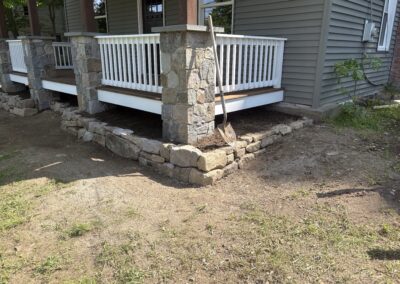 Stone retaining wall under a wooden porch with white railings at a residential property, featuring landscaping work in progress with a shovel resting against the stone pillar, showcasing exterior home improvement and property management.