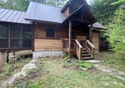 Rustic wooden cabin with a metal roof surrounded by trees and greenery, featuring a small porch with stairs leading to the entrance in a forested property setting.