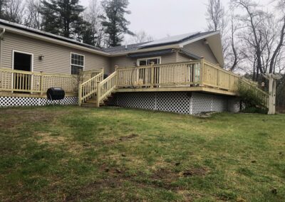Rear view of a single-story beige house featuring a newly constructed wooden deck with lattice skirting, set against a backdrop of leafless trees and green lawn, representing residential property management and home improvement.