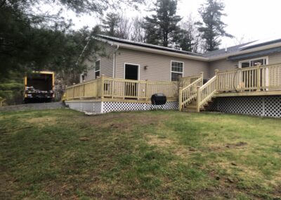 Rear view of a beige single-story home featuring a newly constructed wooden deck with railing and lattice skirting, surrounded by green lawn and trees, illustrating property management and home improvement services.