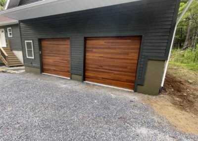 Two modern wooden garage doors set in a dark gray exterior wall of a residential property with gravel driveway and forested surroundings, showcasing quality craftsmanship and exterior home improvement.