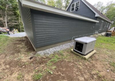 Exterior side view of a modern gray house with fresh landscaping work, including gravel foundation drainage and a newly installed backup generator on a wooden platform, showcasing property management and home energy solutions.